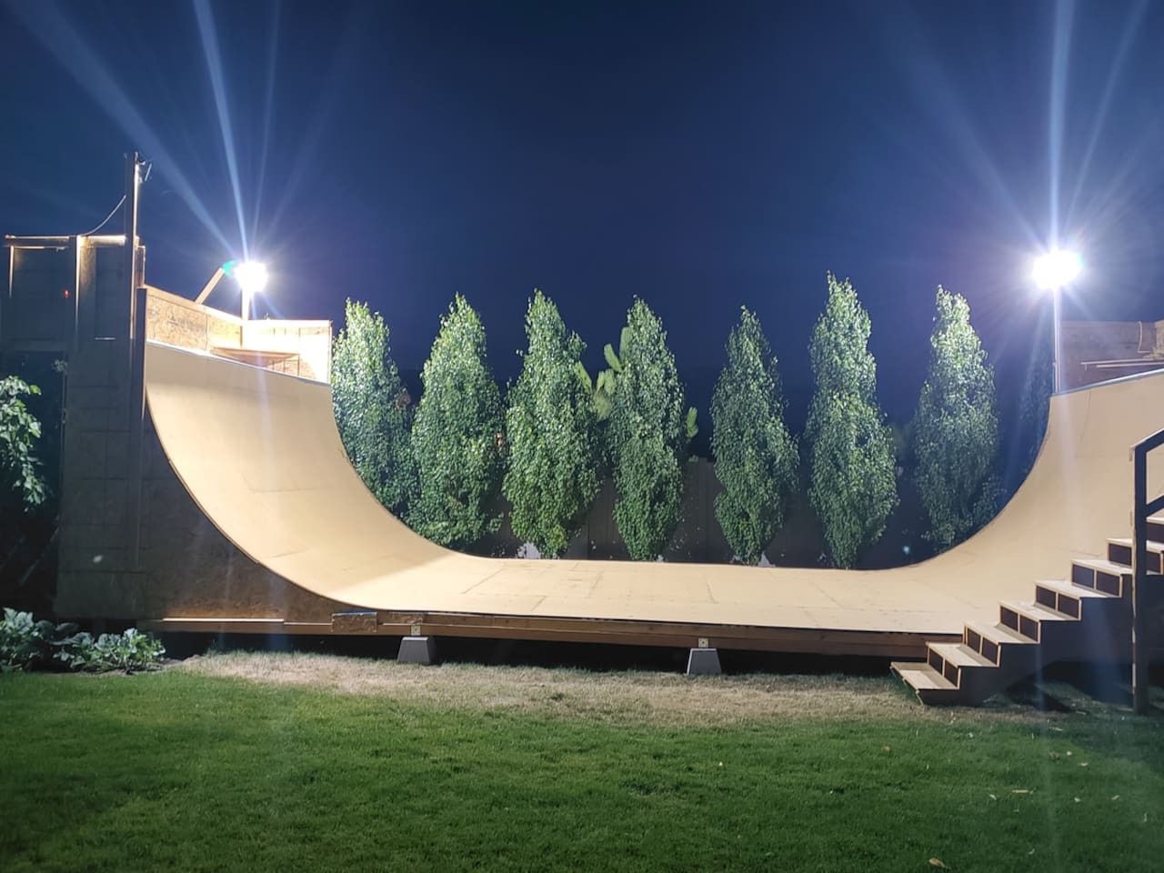 A skatepark with a curved ramp and stairs, surrounded by trees, under artificial lighting.