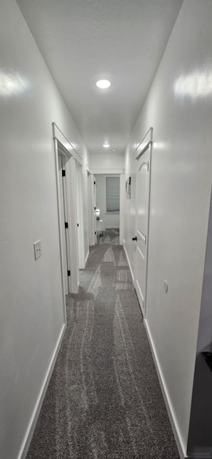 A narrow hallway with carpeted floors and white walls, leading to a room with a window and a door. - Lehi Oasis | Hot Tub | Firepit | 3D Theatre Room (Lehi)