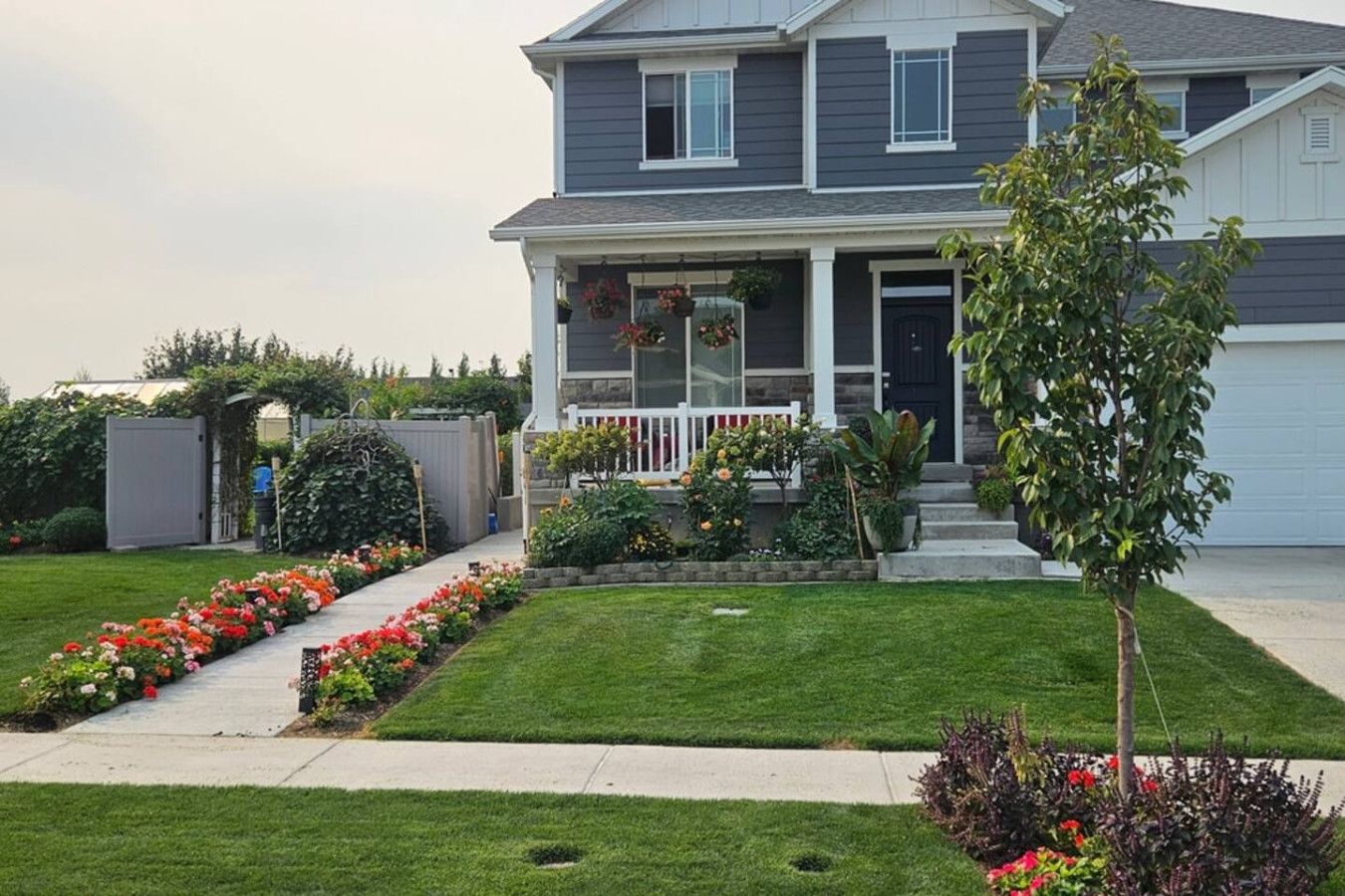 A two-story residential house with a front porch, a small garden with flowers, and a tree.