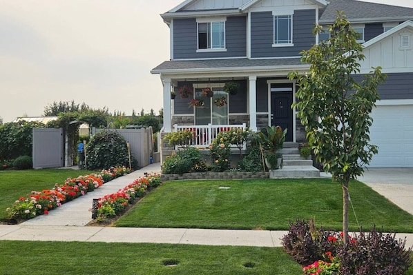 A two-story residential house with a front porch, a small garden with flowers, and a tree.