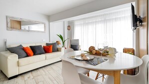 Una sala moderna con un sofá blanco, una mesa de comedor y sillas. Hay un televisor montado en la pared y una ventana grande con cortinas translúcidas.