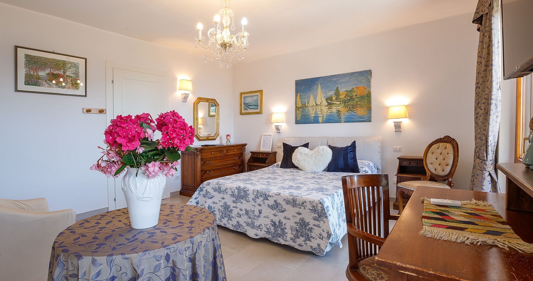 A bedroom with a bed, bedside tables, a chair, a table, a vase with flowers, and wall art.