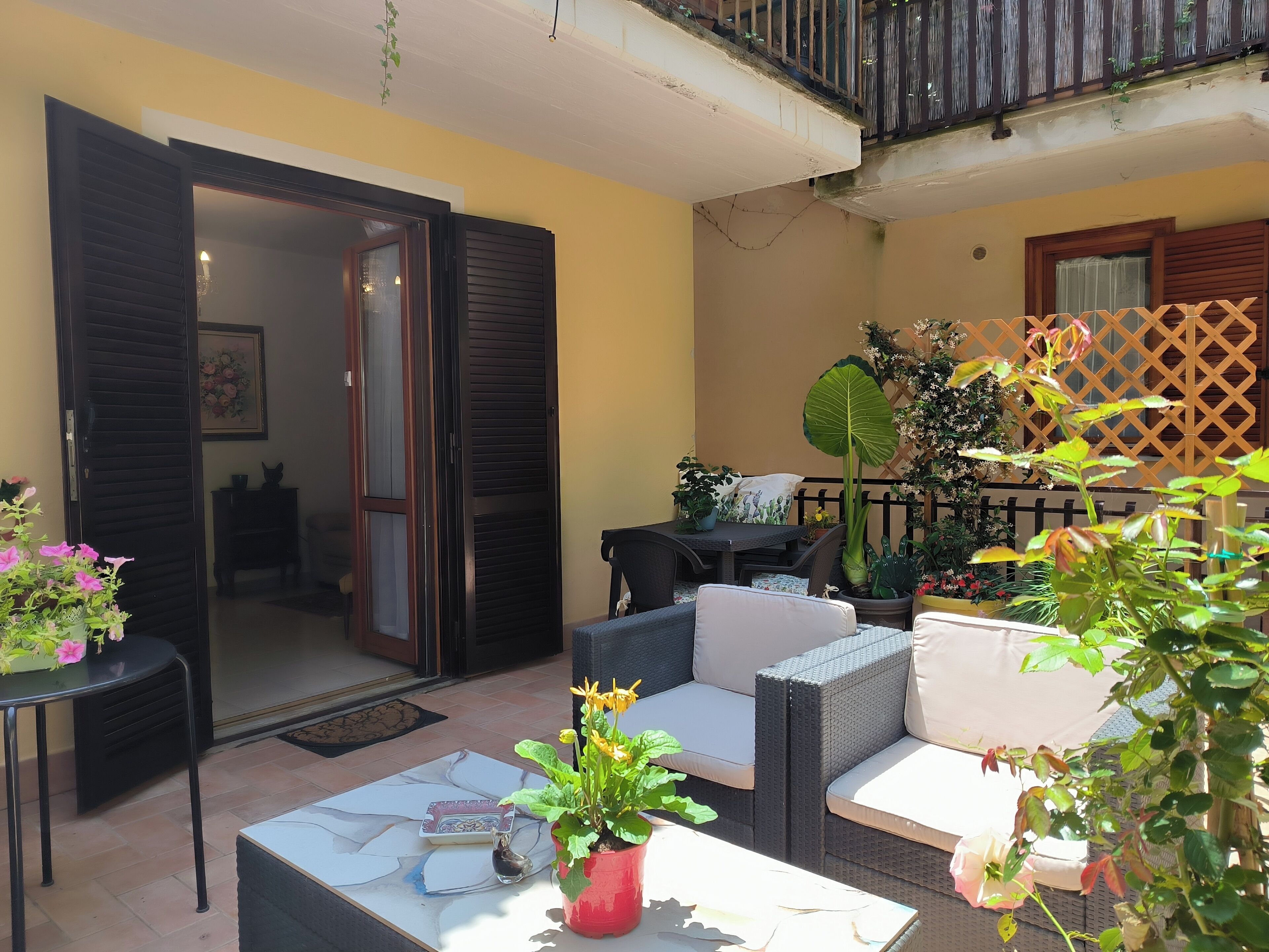 An outdoor patio area with a dining table, chairs, and a sofa. There are potted plants and a large umbrella providing shade.