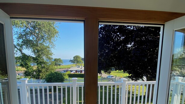 Ein Zimmer mit Holzboden, weißen Geländern und Blick auf eine Terrasse mit weißem Zaun und viel Grün.