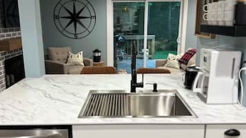 A modern kitchen with a marble countertop, stainless steel sink, and white cabinets. There are two pendant lights, a compass wall decoration, and a view of the outdoors through a sliding glass door.