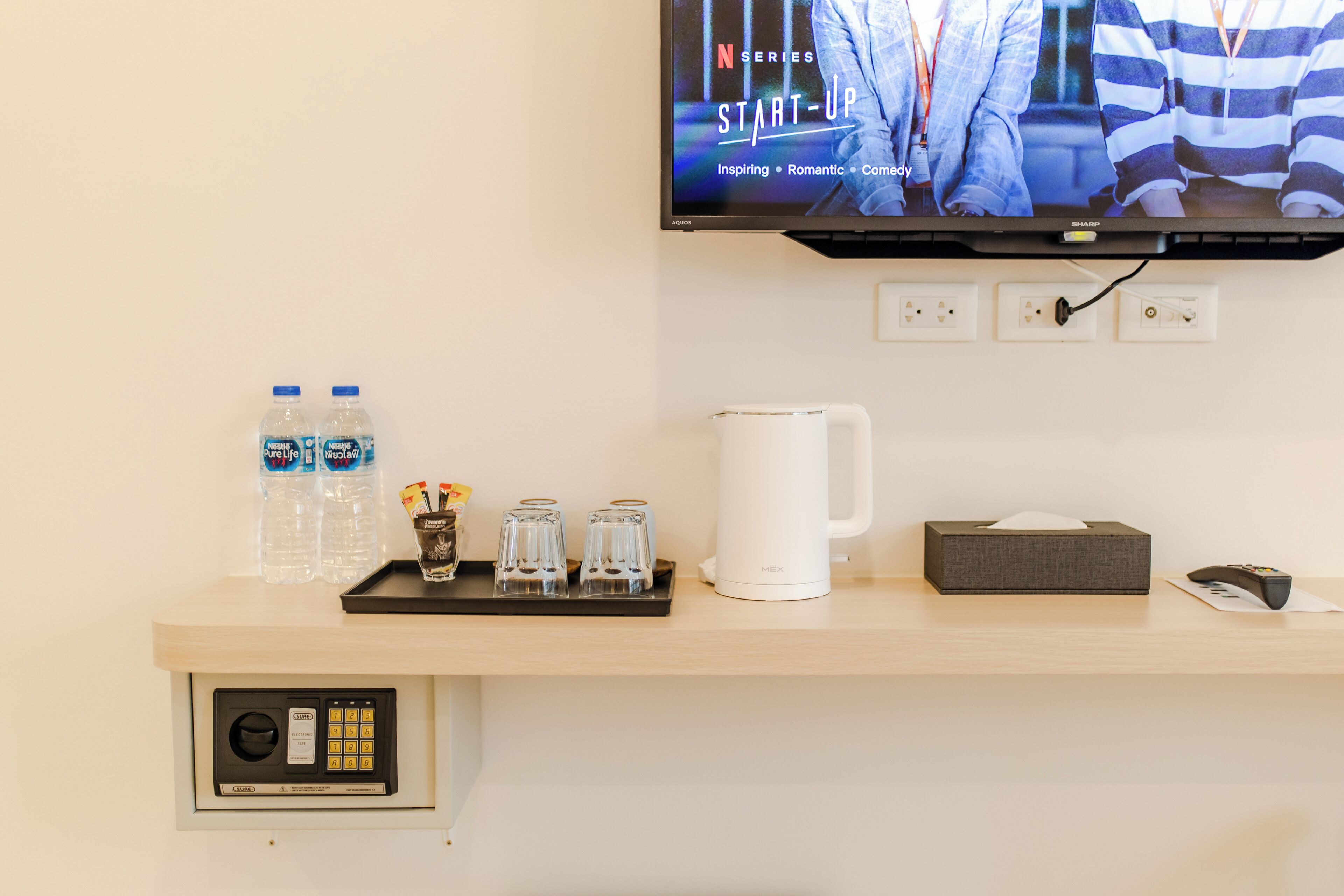 Habitación de hotel con una televisión montada que muestra una serie de Netflix, un minibar con botellas de agua, vasos, una cafetera, una caja de pañuelos y un control remoto.