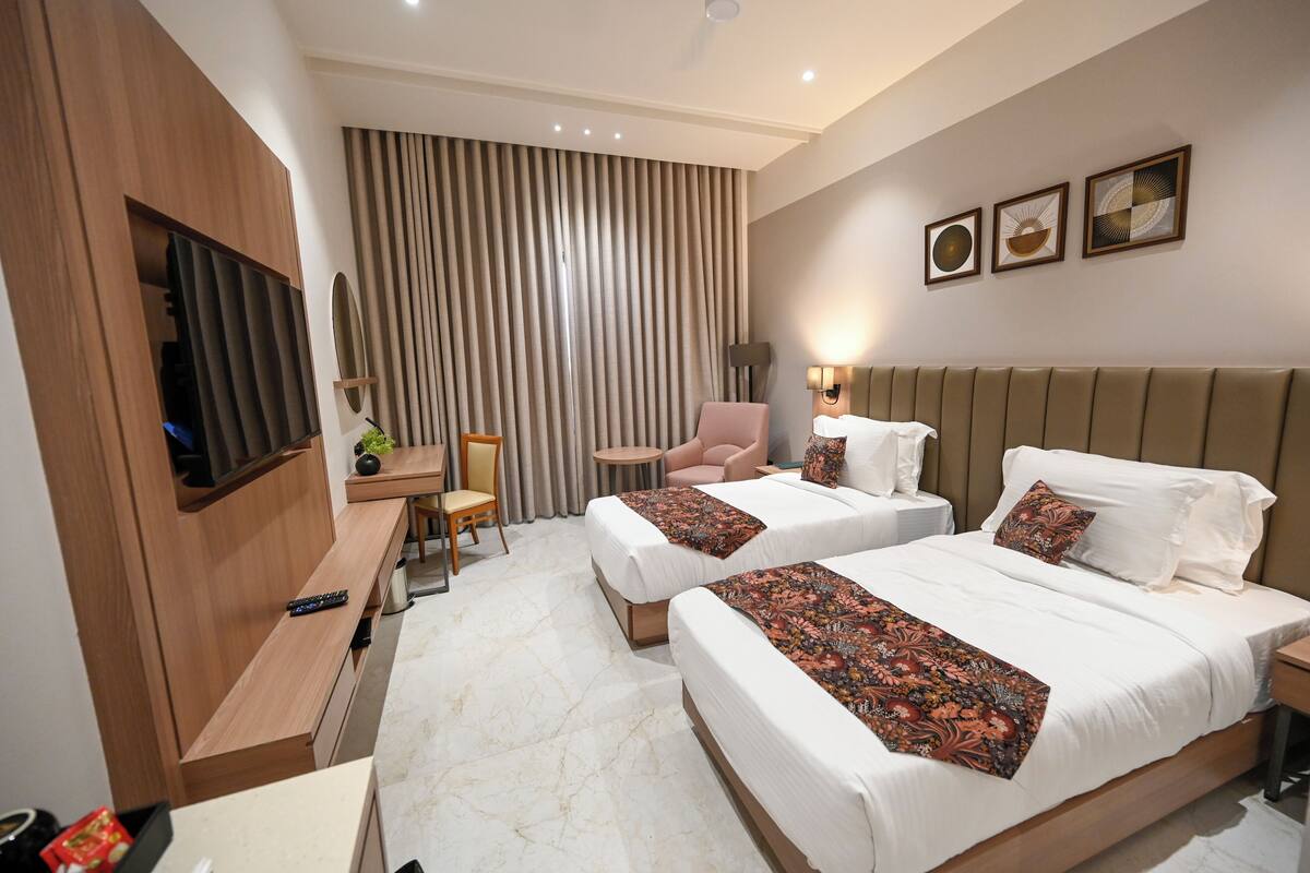 A hotel room with two beds, a desk, a chair, a TV, and a large window with curtains.