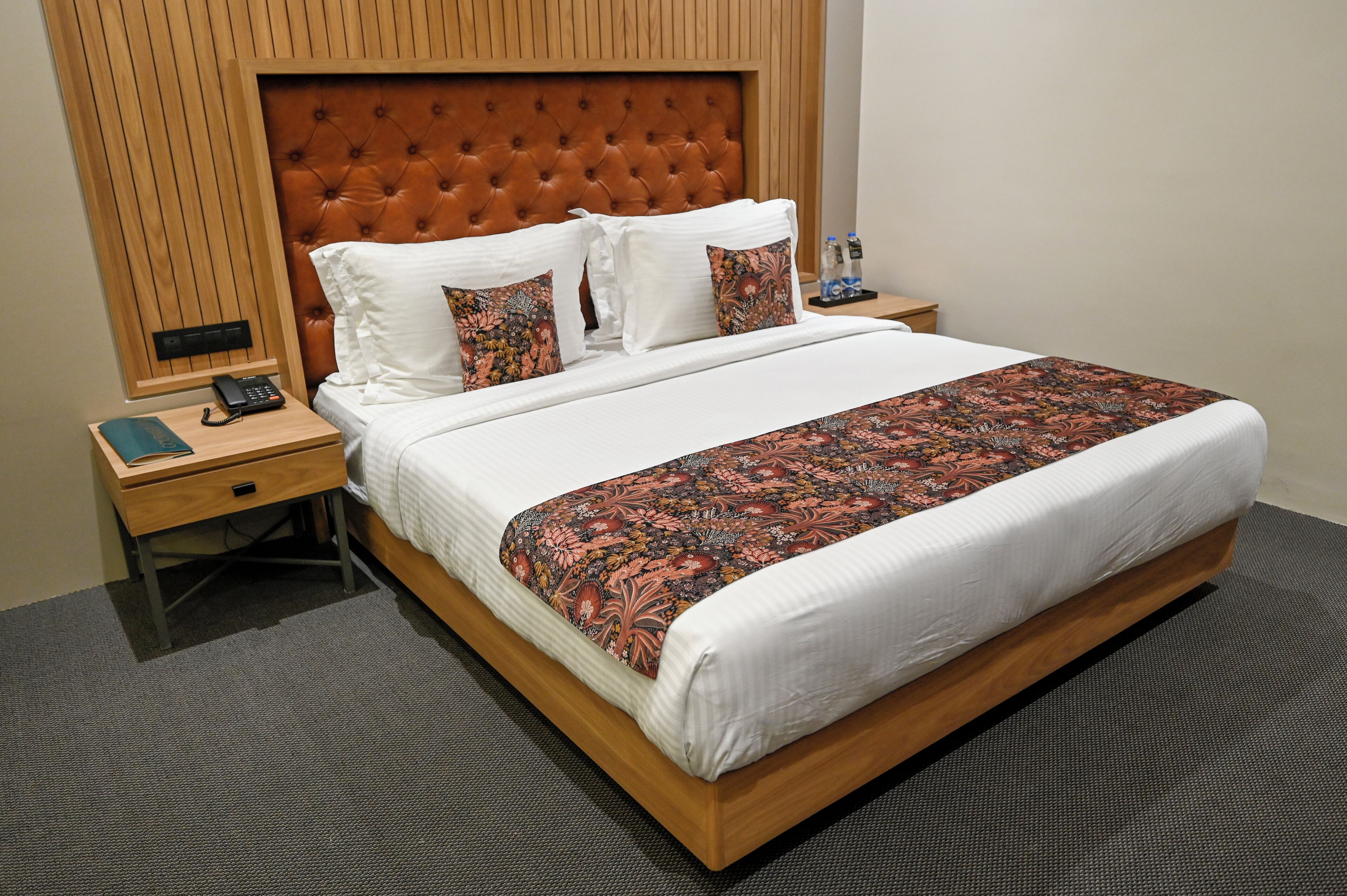 A neatly made bed with a patterned bedspread, a wooden headboard, and two bedside tables with a phone and a bottle.