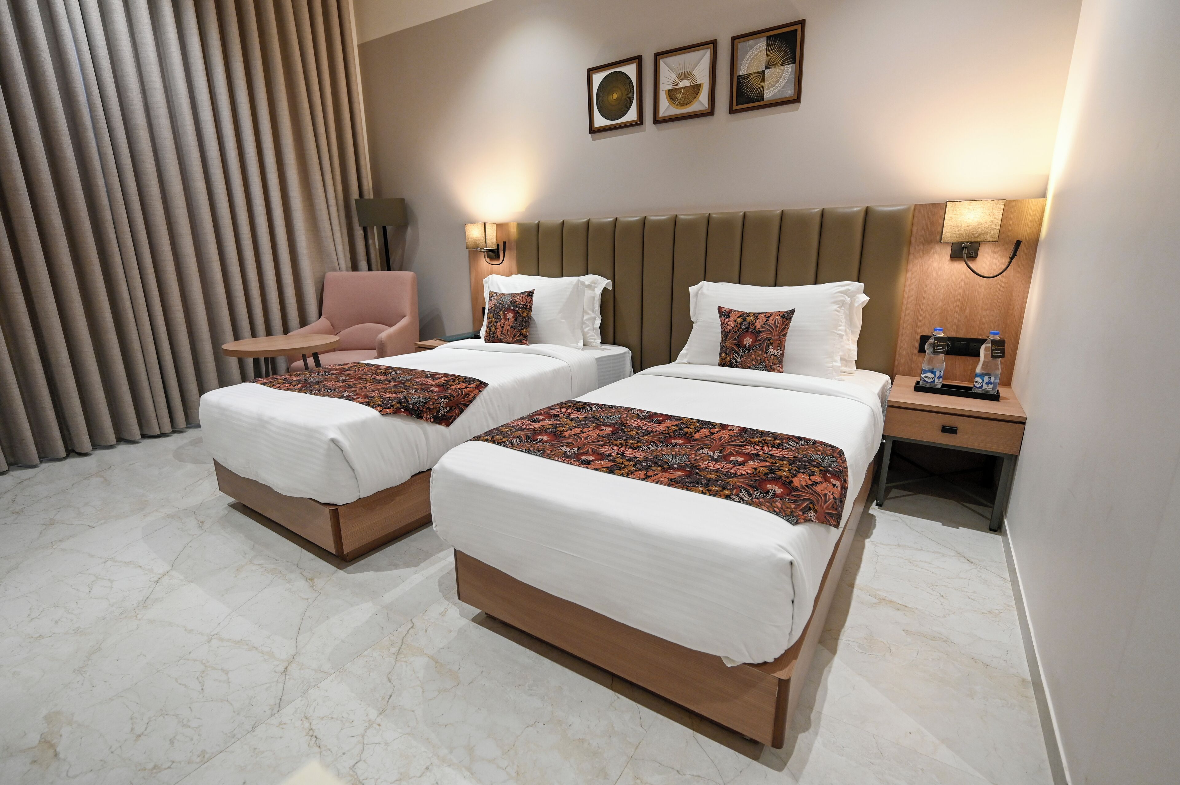 A hotel room with two beds, a chair, a nightstand with a lamp, and a wall-mounted artwork.