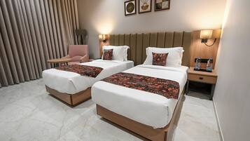 A hotel room with two beds, a chair, a nightstand with a lamp, and a wall-mounted artwork.