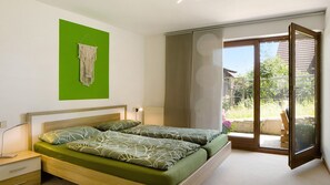 Ein Schlafzimmer mit einem Bett, einem Nachttisch, einer grünen Wand, einer Makramee-Dekoration und einer Glastür, die zu einem Außenbereich führt.