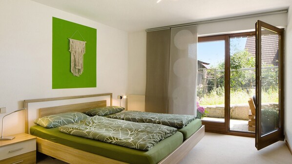 A bedroom with a bed, a bedside table, a green wall, a macrame decoration, and a sliding glass door leading to an outdoor area. - Apartment 'Bäuerle' with Private Terrace, Private Garden and Wi-Fi (Schönwald)