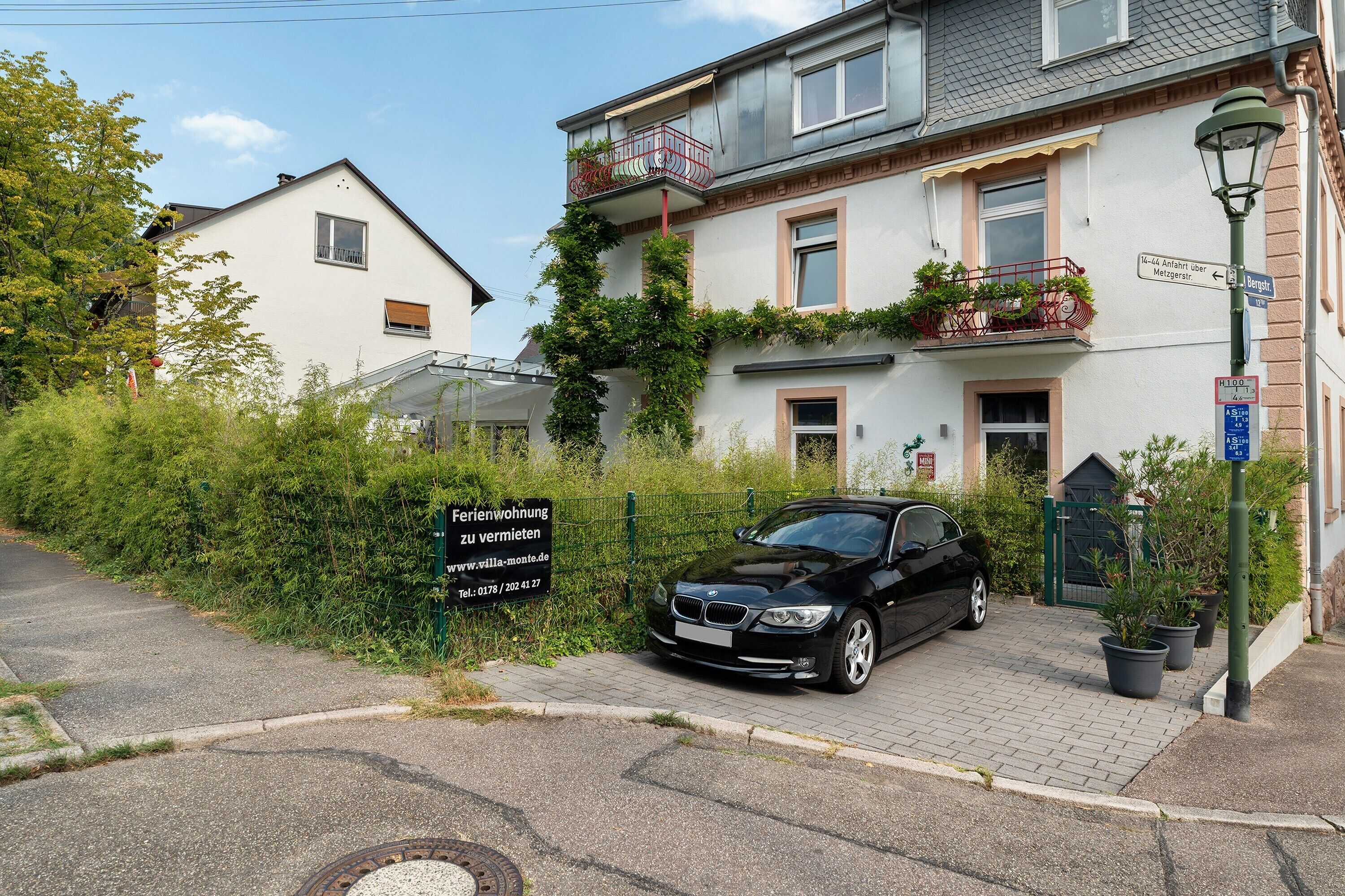 A residential building with a black car parked in front, a sign indicating a property for sale, and a street lamp.