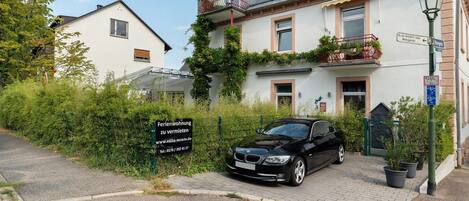 A residential building with a black car parked in front, a sign indicating a property for sale, and a street lamp.