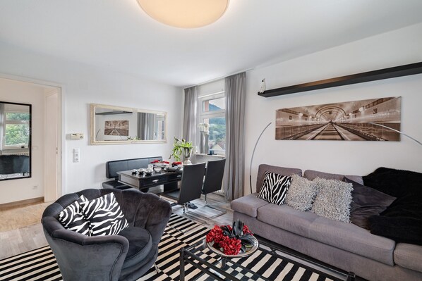A modern living room with a sofa, armchair, dining table, and a large painting on the wall.