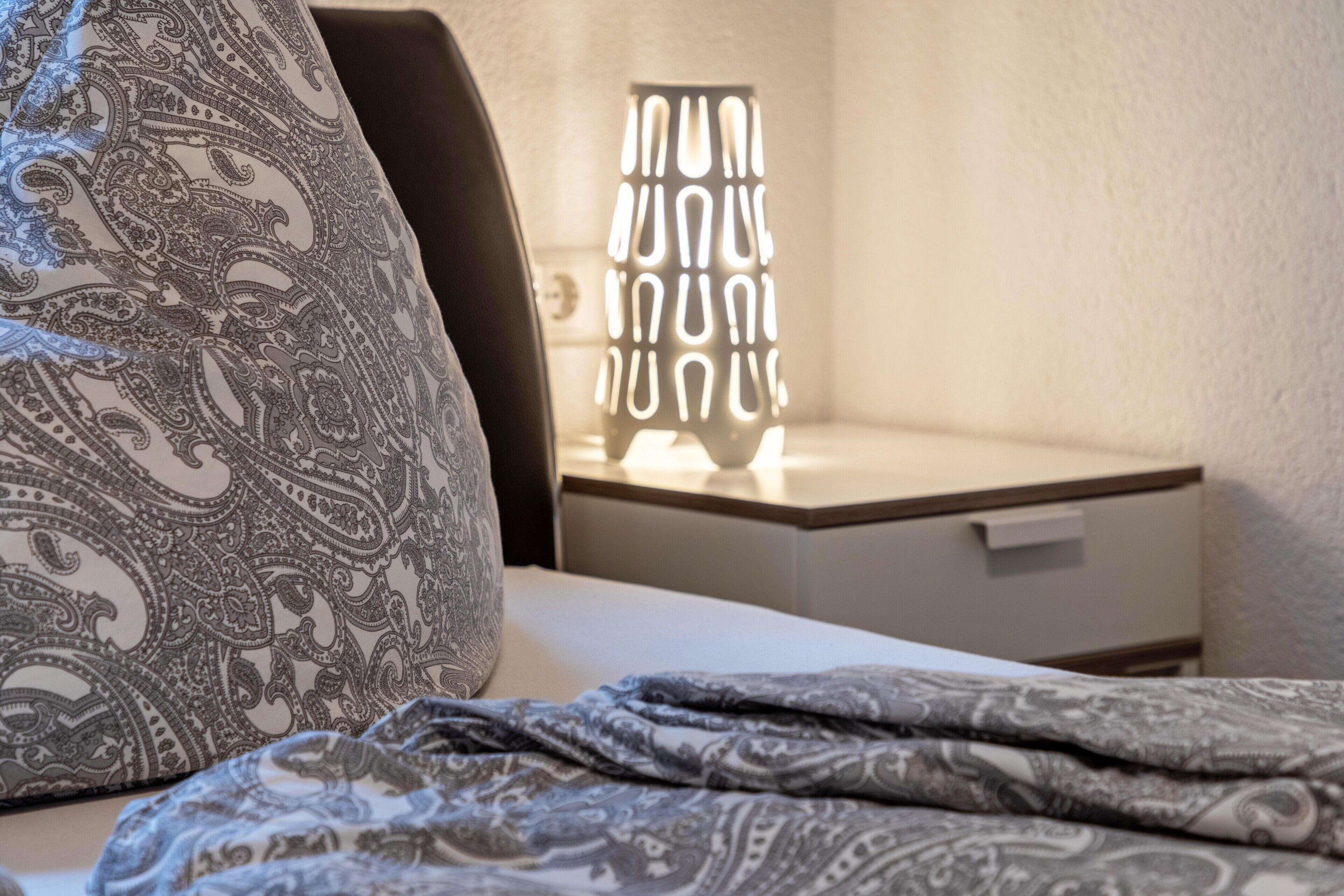 En säng med mönstrat sänglinne, en sänglampa och ett nattduksbord med en låda.