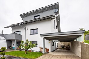 Une maison moderne à deux étages, dotée d’un patio couvert, d’une antenne parabolique et d’une petite pelouse.