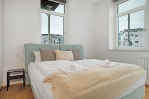 Een slaapkamer met een bed, kussens, een nachtkastje en een raam met uitzicht op gebouwen.