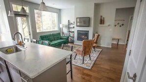 A modern kitchen with a large island, a dining area with a green sofa and a fireplace, and a wooden floor. - Plumberry Cottage in Charming Neighborhood (College Place)