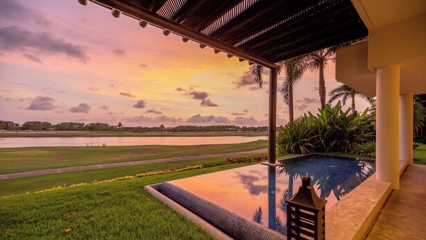 Una piscina con vista a un atardecer sobre una masa de agua, rodeada de follaje y palmeras.