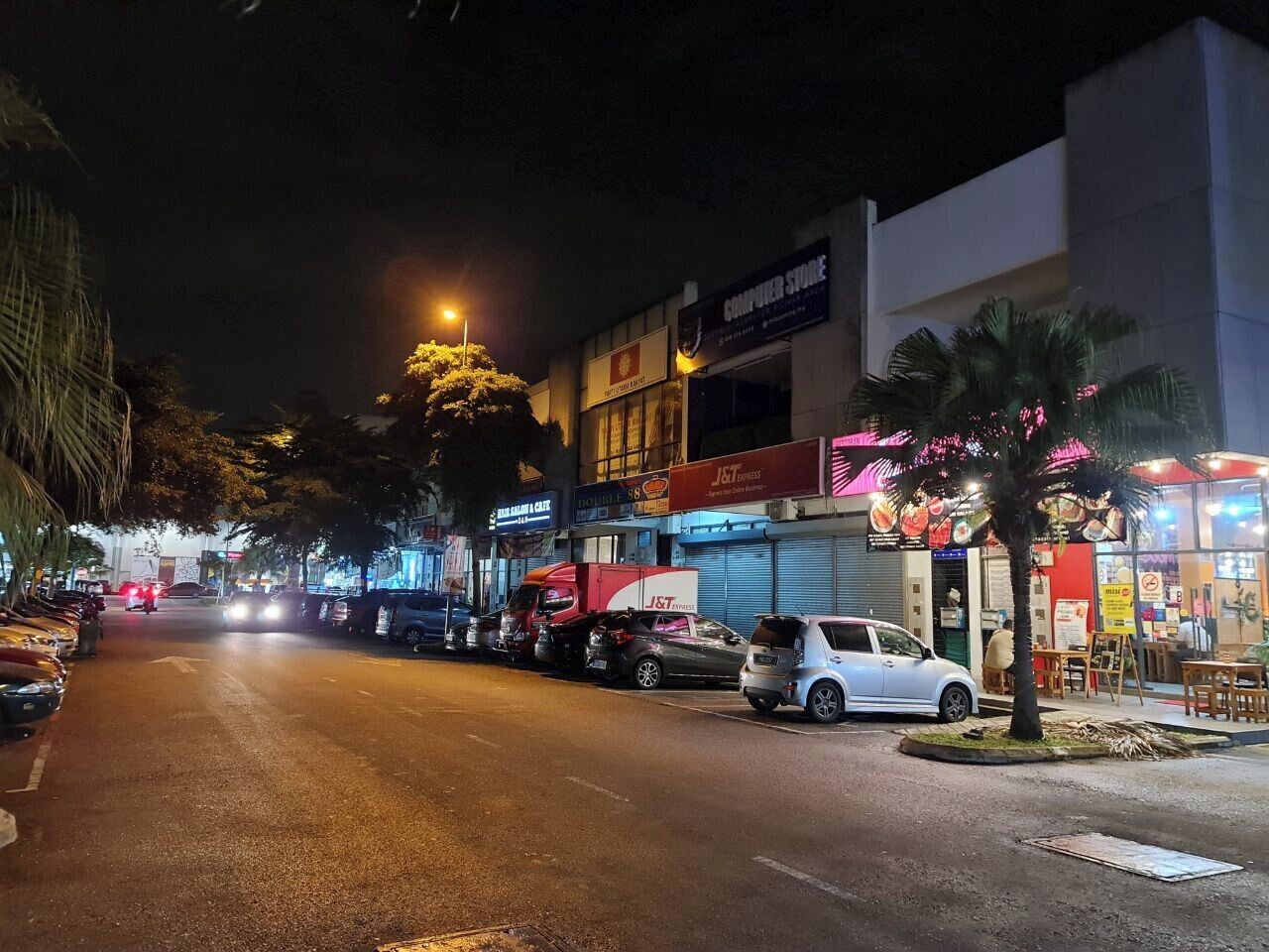 A nighttime street scene with parked cars, a J&T Express store, and a computer store.