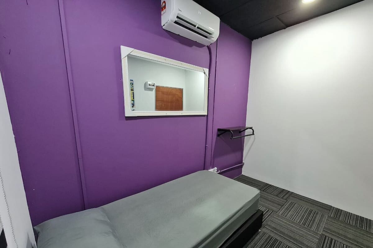 A small, single-bed room with a purple wall, a window with a wooden panel, and a white air conditioning unit.