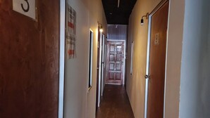 Um corredor estreito com piso e paredes de madeira, que leva a um quarto com porta de madeira. Há placas com números de quarto e um banheiro.