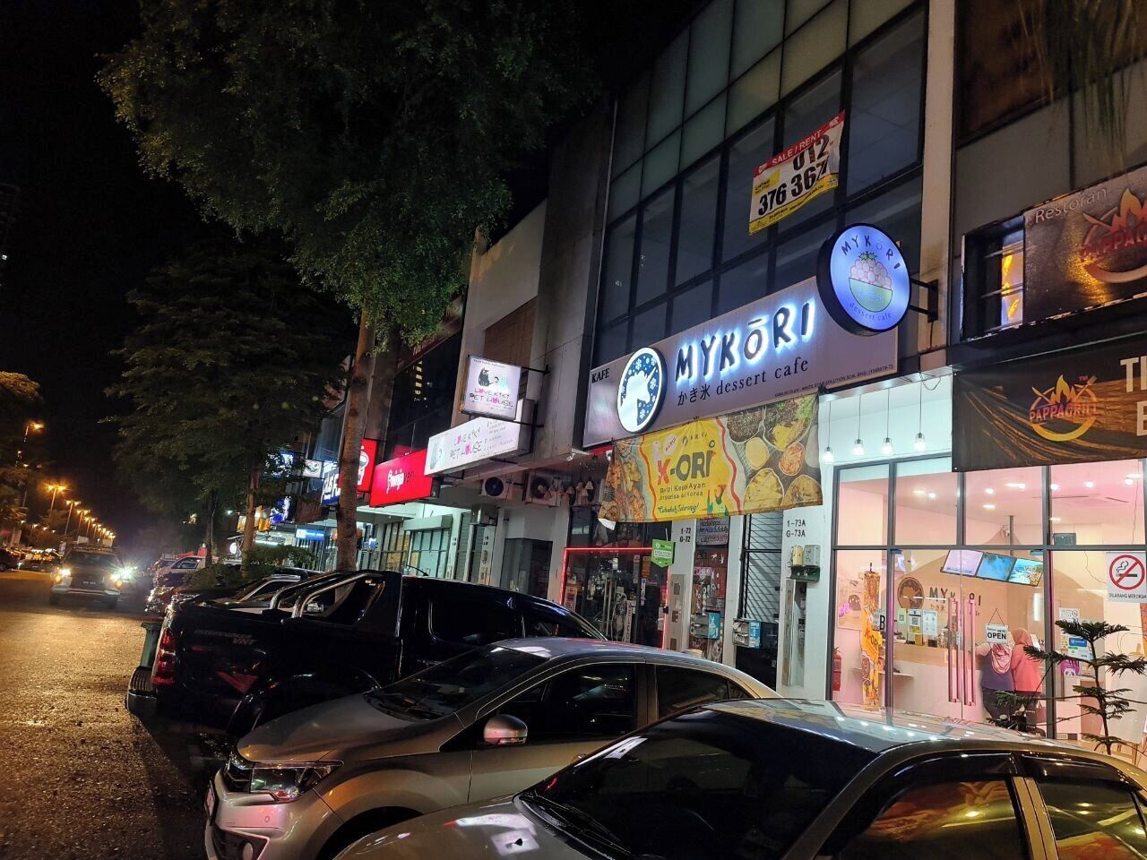 A street view at night with parked cars and illuminated storefronts, including a cafe named MYKORI.