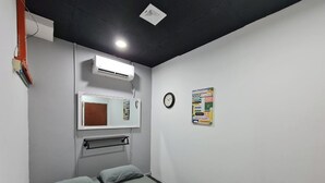 Quarto pequeno com uma cama de solteiro, colchão cinza, ar condicionado branco, espelho fixado na parede, relógio e painel de avisos.