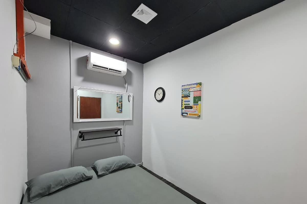A small, single-bed room with a grey mattress, a white air conditioning unit, a wall-mounted mirror, a clock, and a bulletin board.