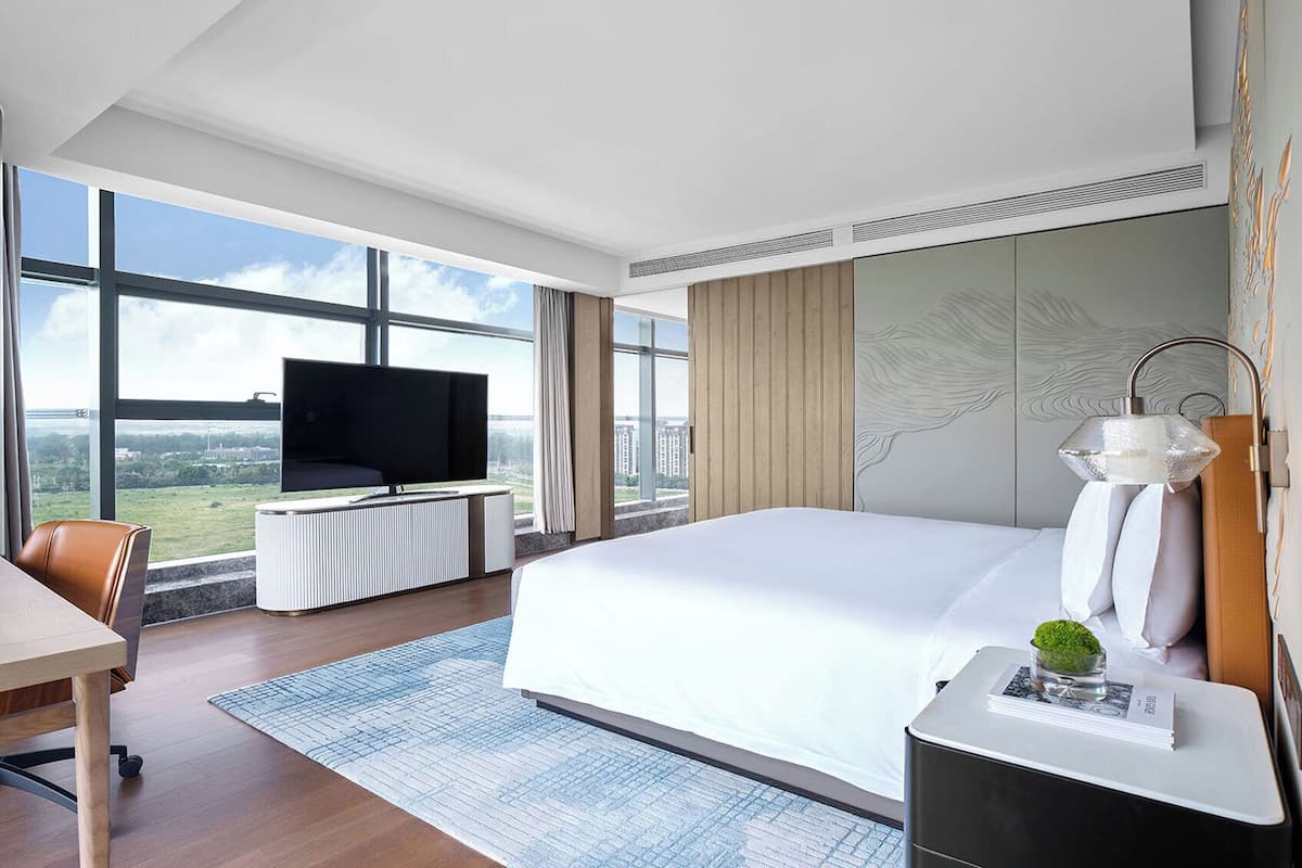 A modern hotel room with a large bed, a flat-screen TV, a desk, and a view of the cityscape.