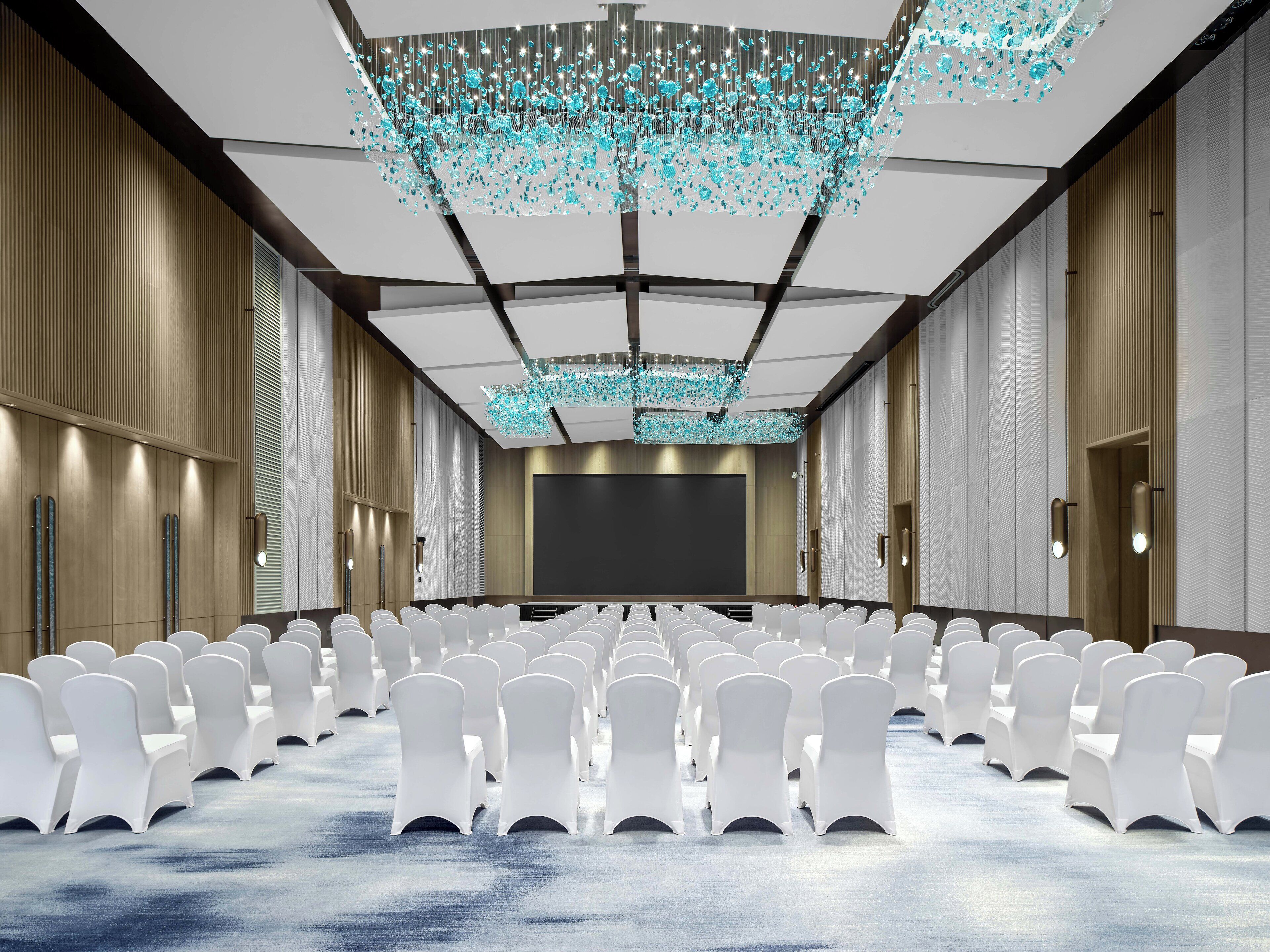 Une salle de conférence avec des rangées de chaises blanches, un grand écran et un plafond orné de luminaires suspendus.