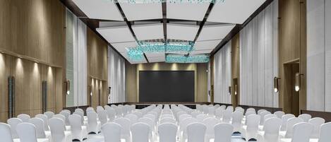 Une salle de conférence avec des rangées de chaises blanches, un grand écran et un plafond orné de luminaires suspendus.