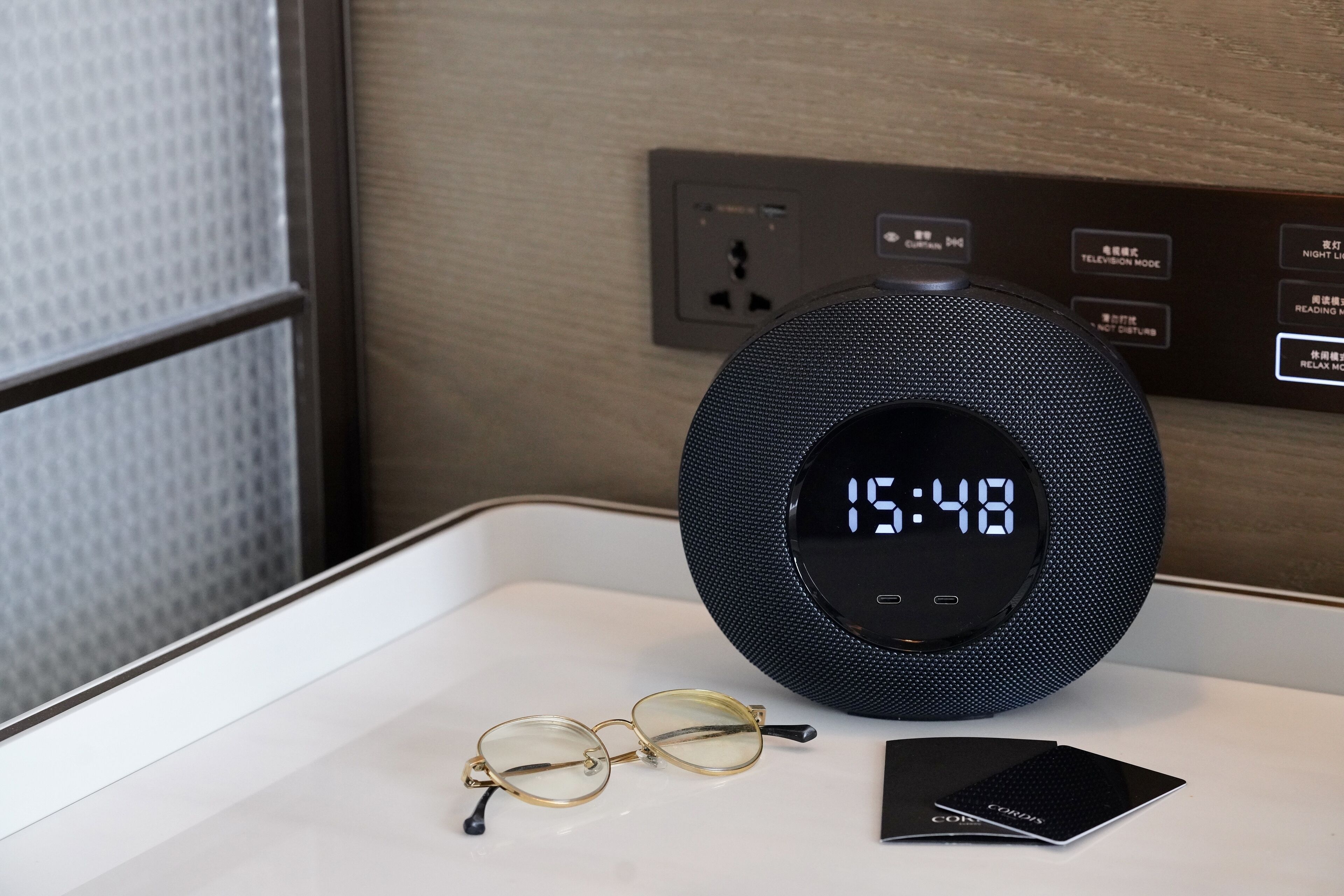 Une horloge numérique affichant 15h48, une paire de lunettes et un carnet noir sur une surface blanche.