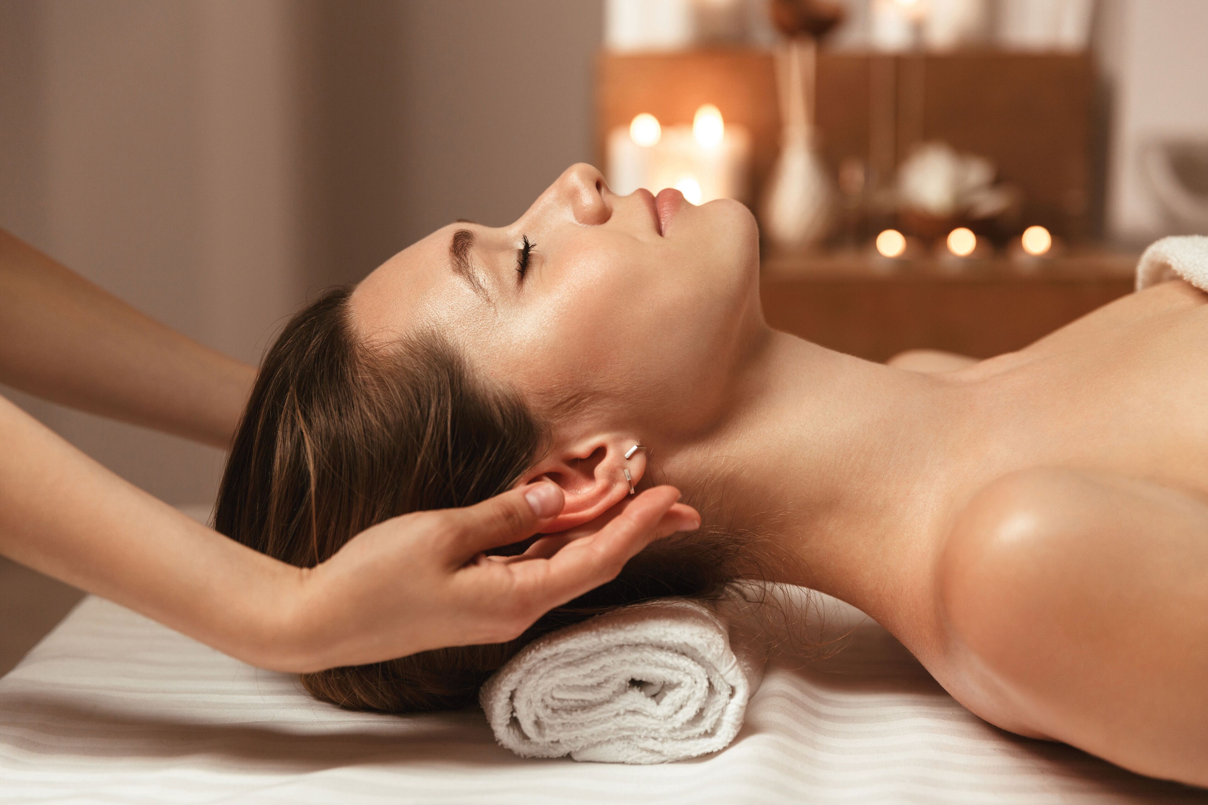 A person lying on a massage table with a towel rolled at the head, in a room with candles in the background.