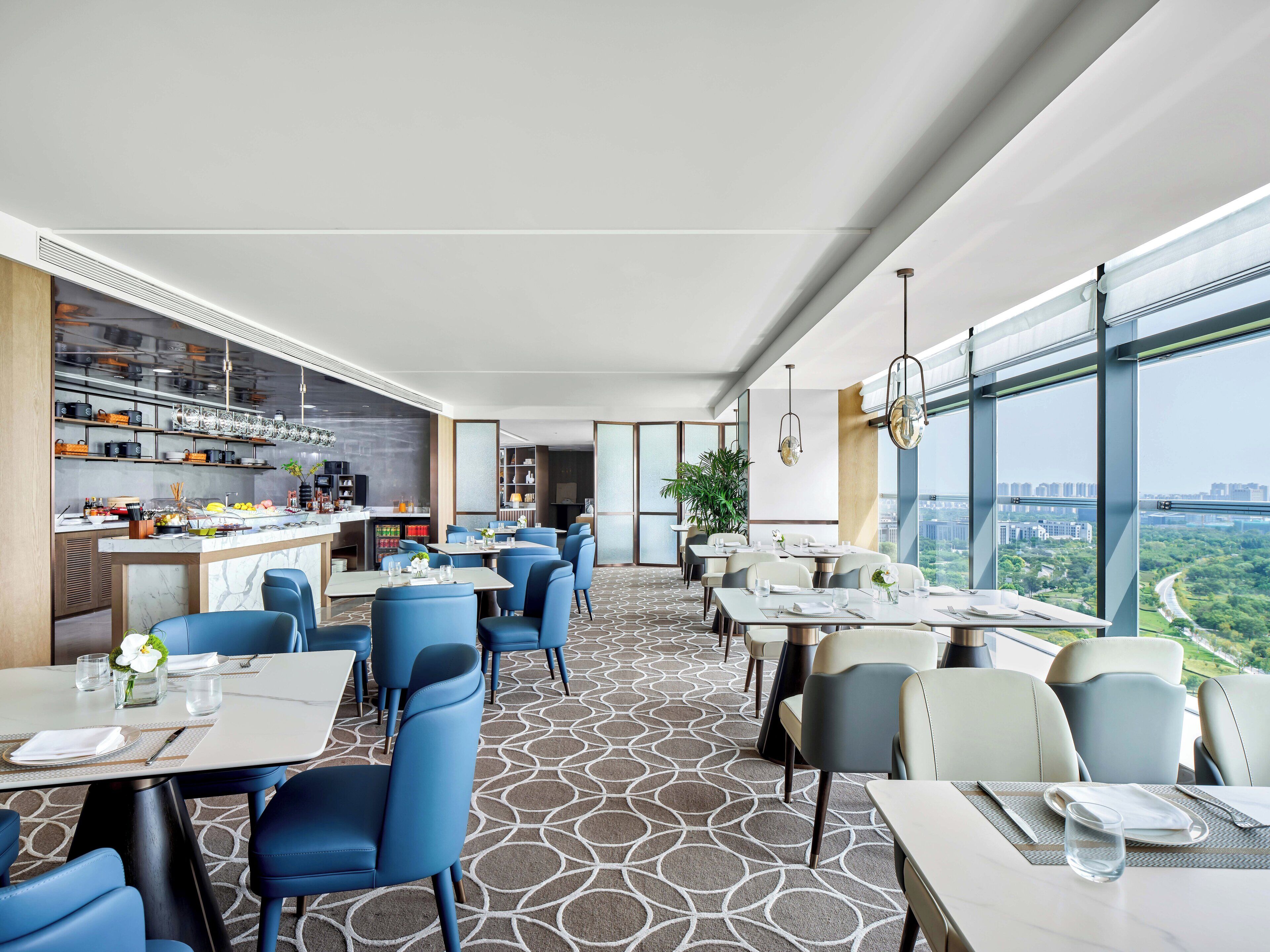 A modern restaurant with a high ceiling, large windows offering a city view, and a central bar area.