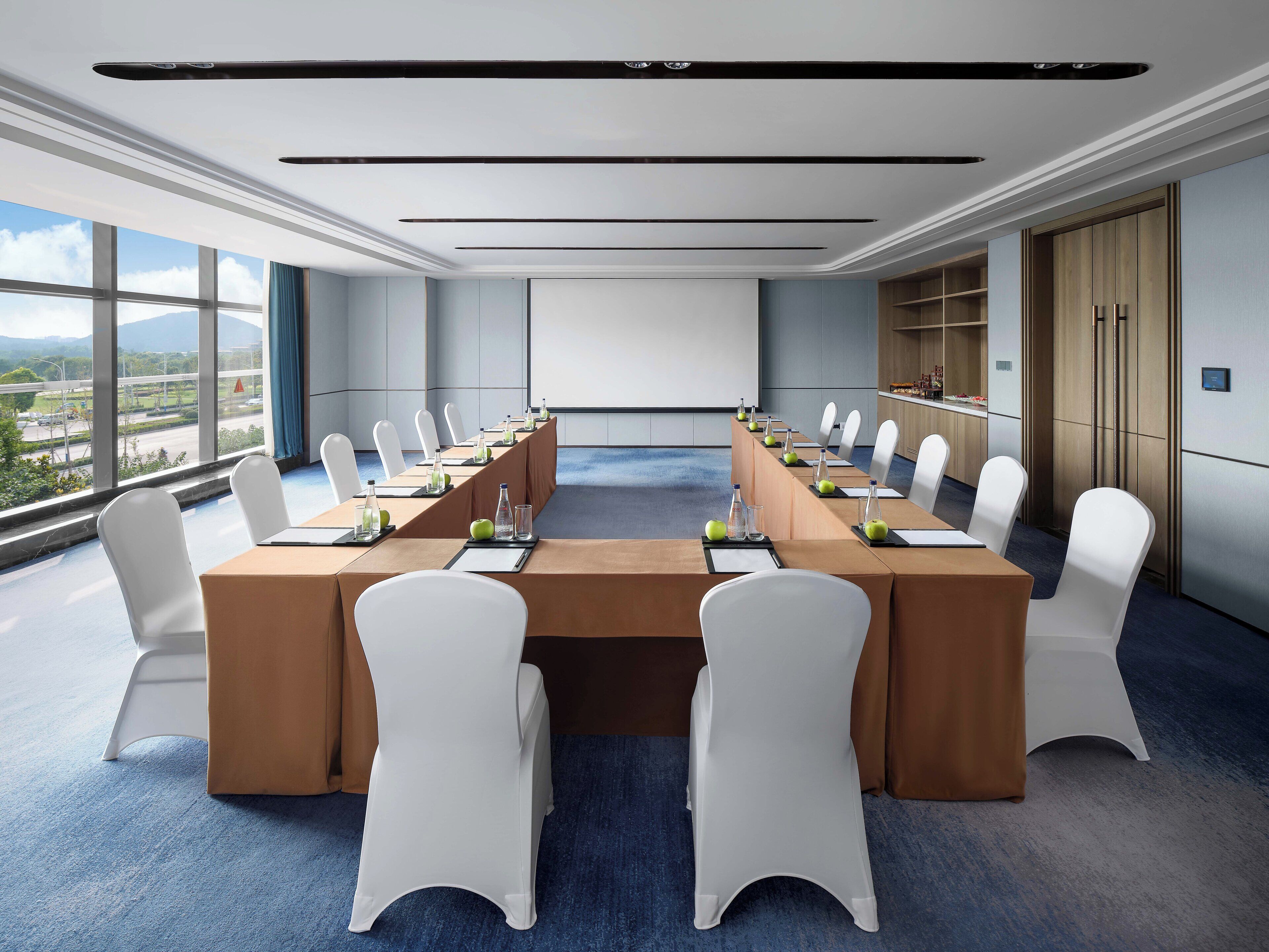 Un salón de conferencias con una larga mesa, sillas blancas y vistas al exterior.