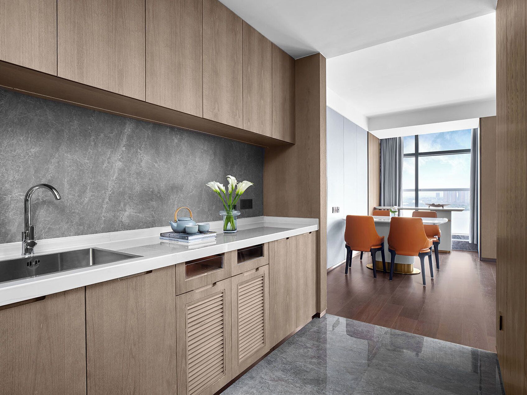 A modern kitchen with wooden cabinets, a white countertop, and a view of a dining area with orange chairs.