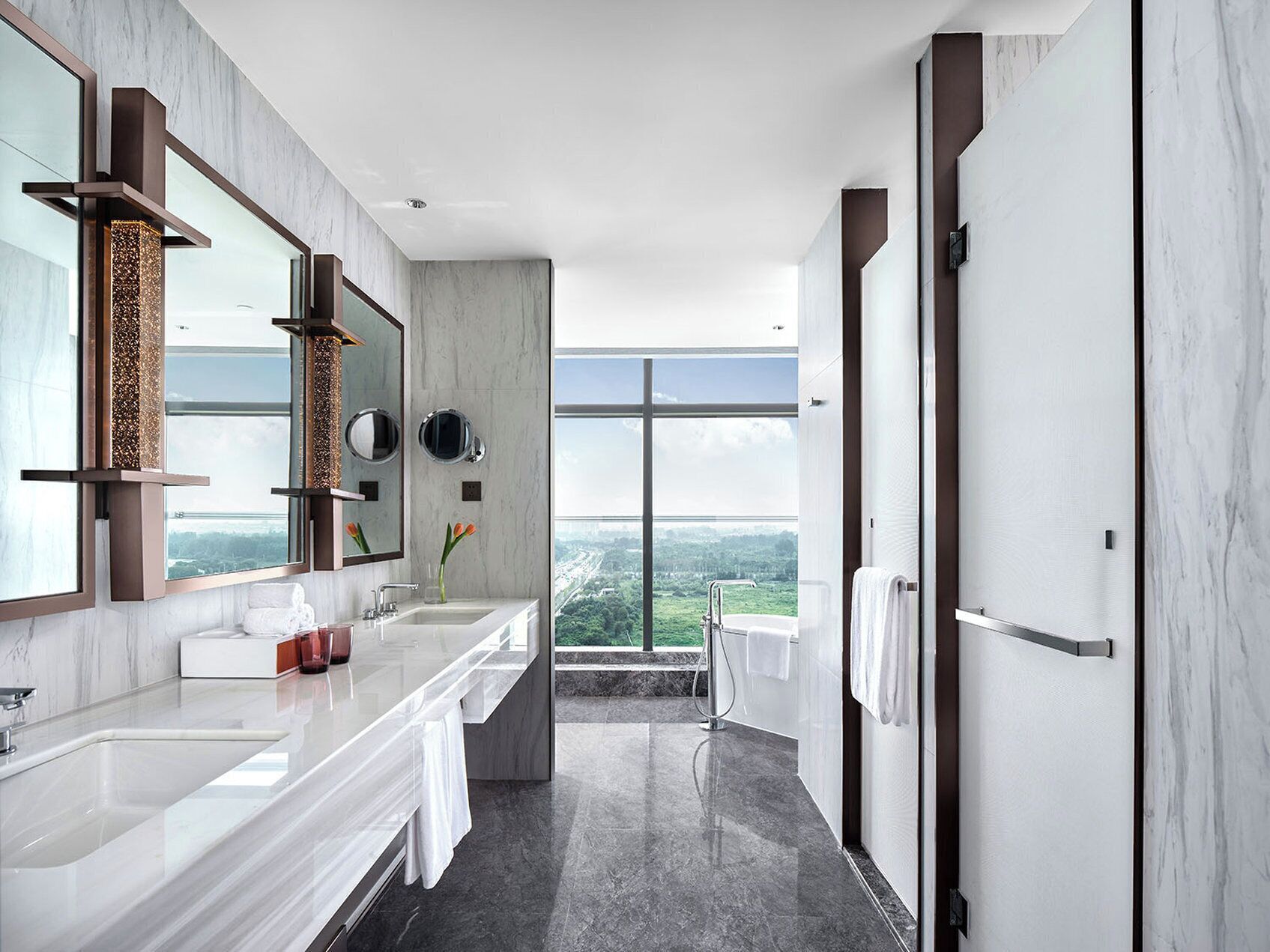 A modern bathroom with a large mirror, two sinks, a shower, and a view of a cityscape.