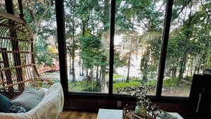 Una habitación acogedora con un ventanal grande, una silla de mimbre, un sofá blanco y una mesita con un jarrón y un libro.