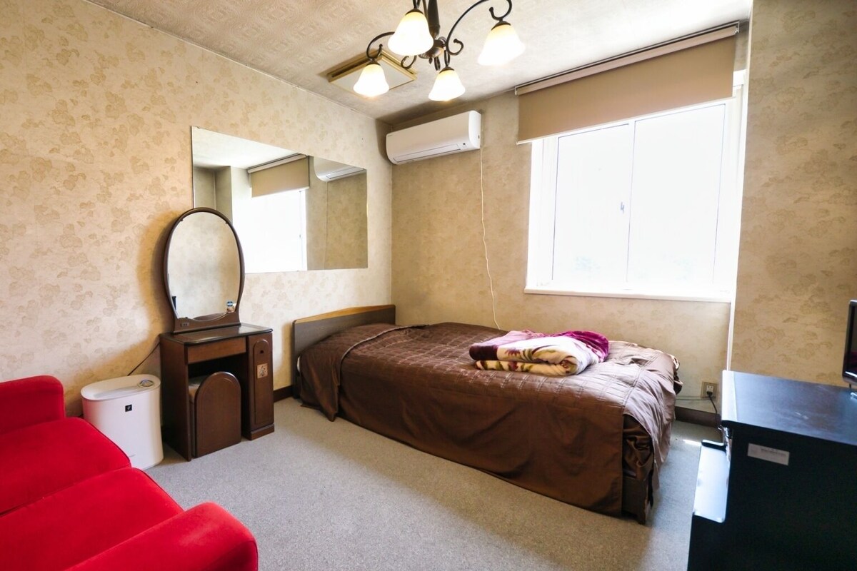 Un dormitorio con cama, una silla roja, un escritorio con espejo y una ventana.