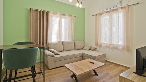 A living room with a green wall, a beige sofa, a wooden coffee table, a TV, and a dining area with a green chair and a table. - Aristea in Athens With 2 Bedrooms and 1 Bathrooms (Athens)