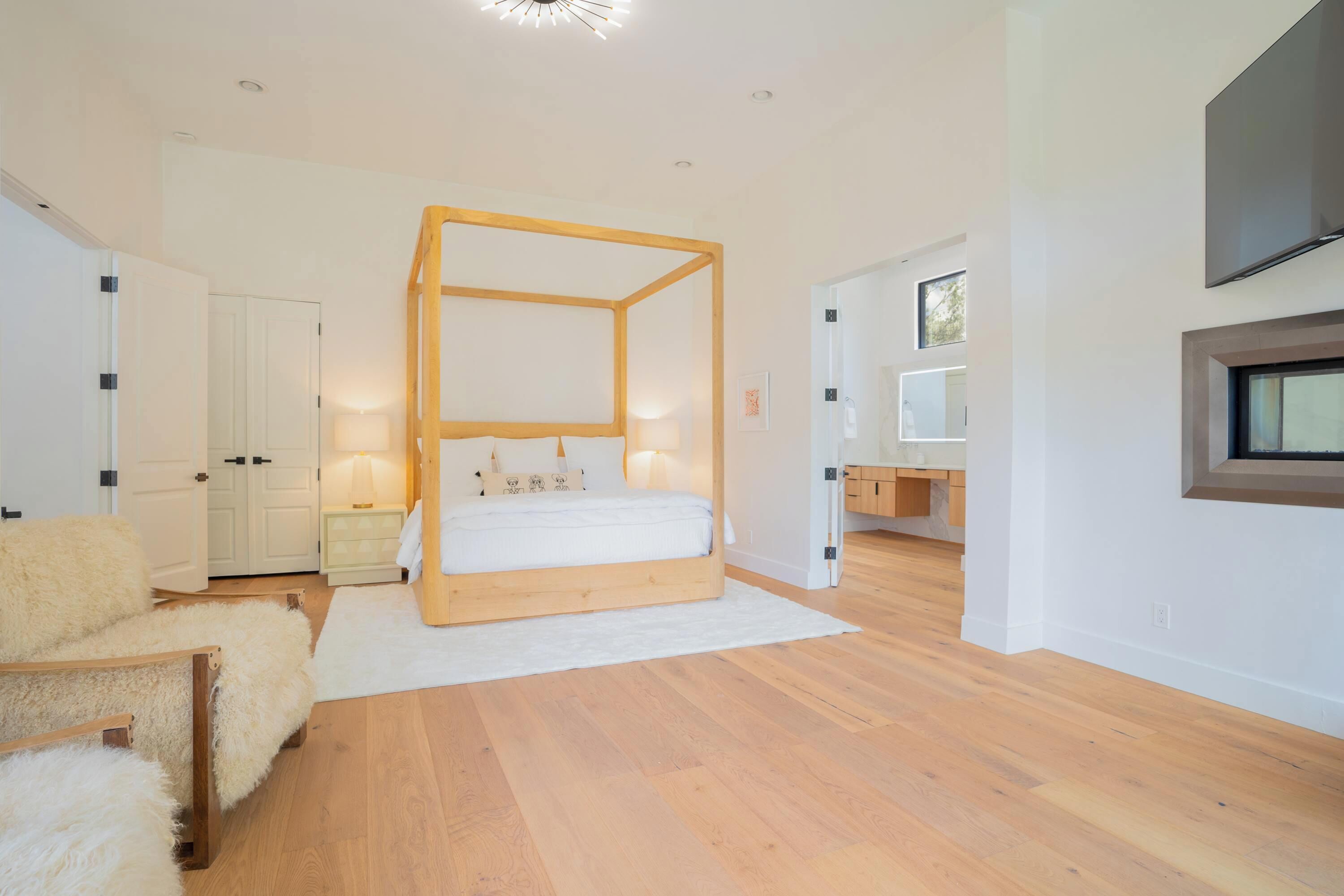 Un dormitorio moderno con una cama con dosel, suelo de madera, un armario blanco, una mesita de noche y un televisor montado en la pared.