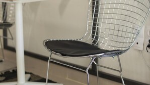 Un interior modern amb una cadira de filferro, una taula blanca i una estant amb objectes decoratius.