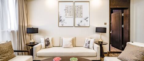 Una sala de estar moderna con un sofá color beige, dos mesitas de noche con lámparas, una mesa de centro con cuencos de fruta y dos piezas de caligrafía enmarcadas en la pared.