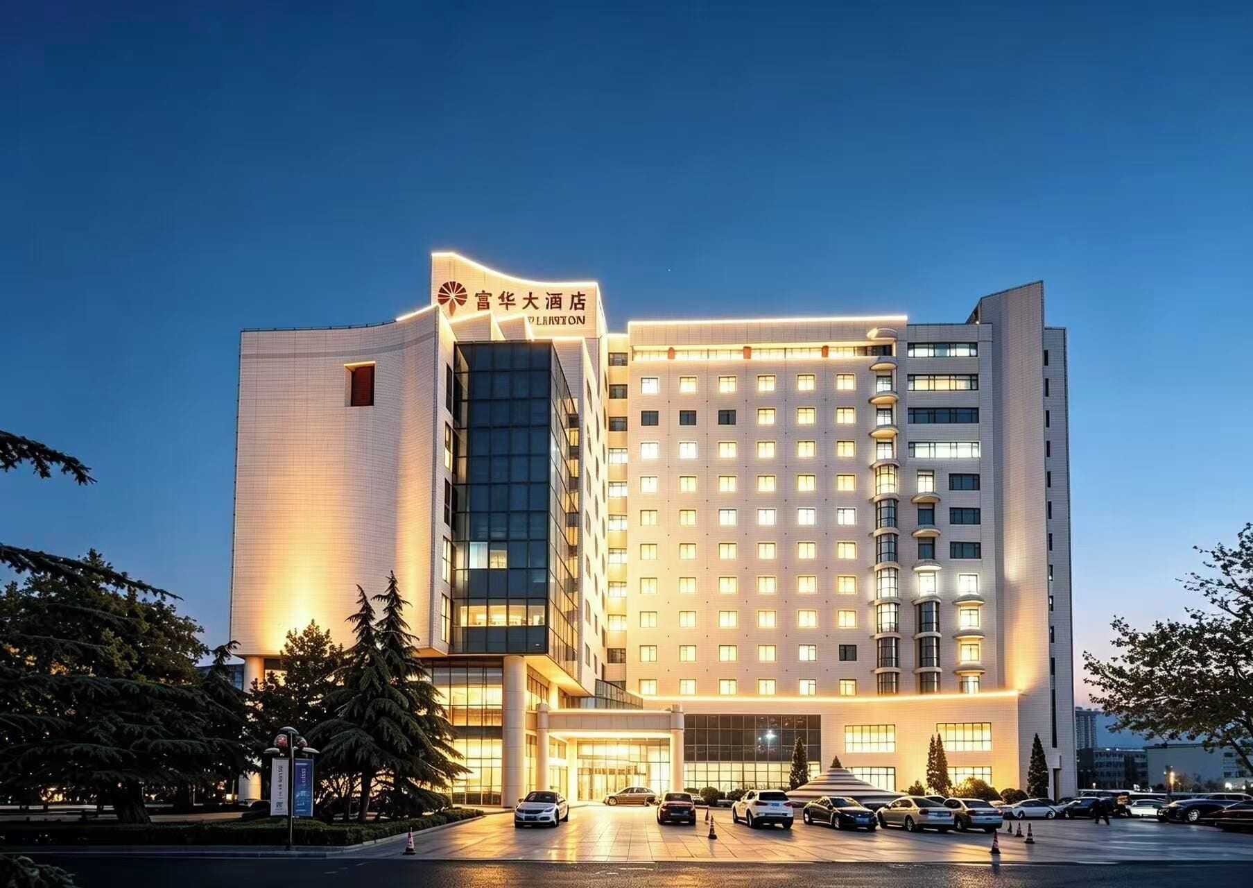 Un hotel de varios pisos con fachada moderna, ventanas iluminadas y un estacionamiento con varios automóviles.