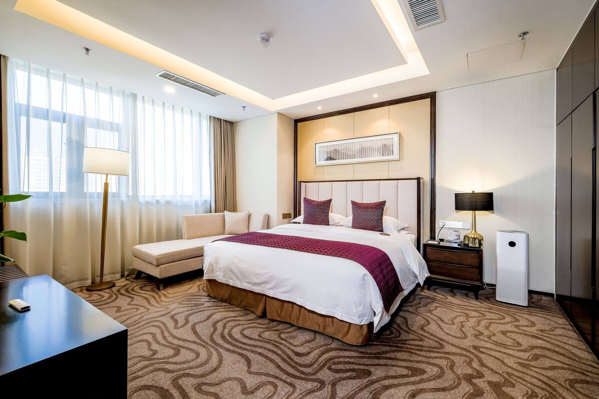 A hotel room with a large bed, a bedside table with a lamp, a desk with a phone, and a large window with curtains.