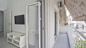 A balcony with a view of buildings, a sliding glass door, a TV mounted on the wall, and a white bench. - Sofia s Home in Piraeus (Piraeus)