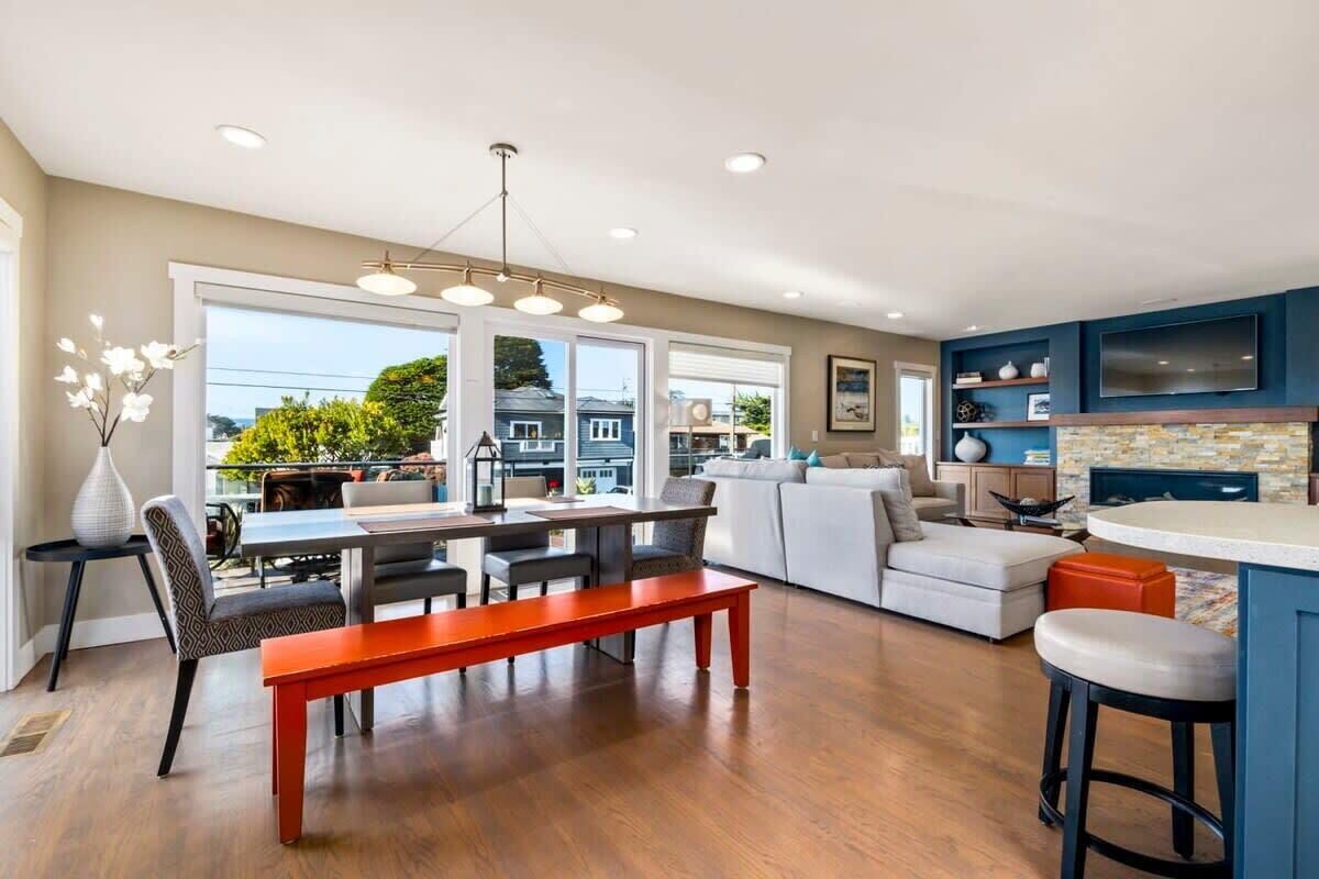 A spacious living area with a dining table, seating, a kitchen with a TV, and large windows.
