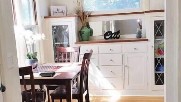Une vue intérieure d’un coin repas avec une table et des chaises en bois, une cuisine en arrière-plan et un paillasson à l’entrée.